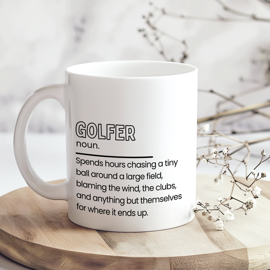 White ceramic mug with “Golfer noun.” definition style design in black typography, featuring a funny quote about chasing a golf ball around a field and blaming everything else, displayed on a neutral wooden background.