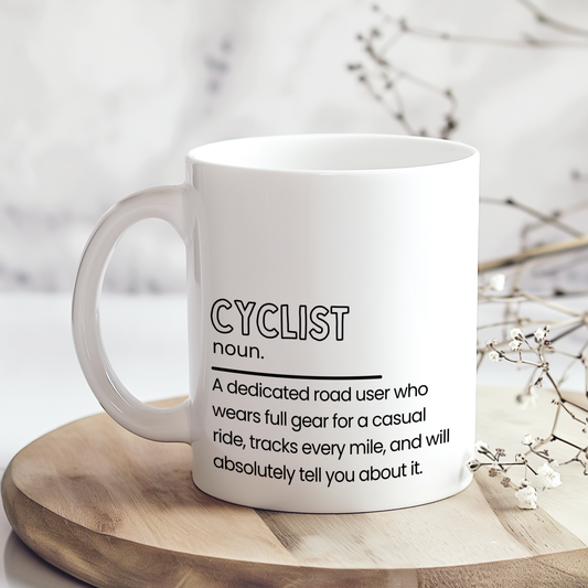 White ceramic mug with “Cyclist noun.” definition style quote in black typography, featuring a funny description about cyclists wearing full gear and tracking every mile, displayed on a neutral wooden background.
