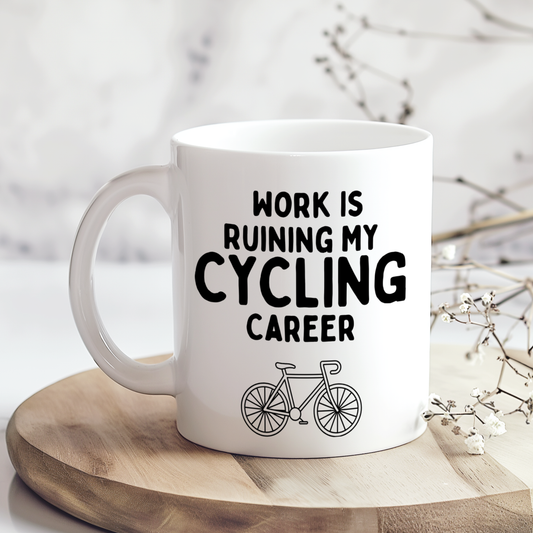White mug with black “Work Is Ruining My Cycling Career” lettering and a road bicycle illustration, displayed on a wooden board beside dried flowers.