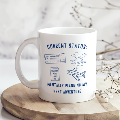 White ceramic mug featuring the phrase "Current Status: Mentally Planning My Next Adventure" with blue illustrations of a boarding pass, passport, aeroplane and sunset. Perfect gift for travel lovers and holiday enthusiasts.