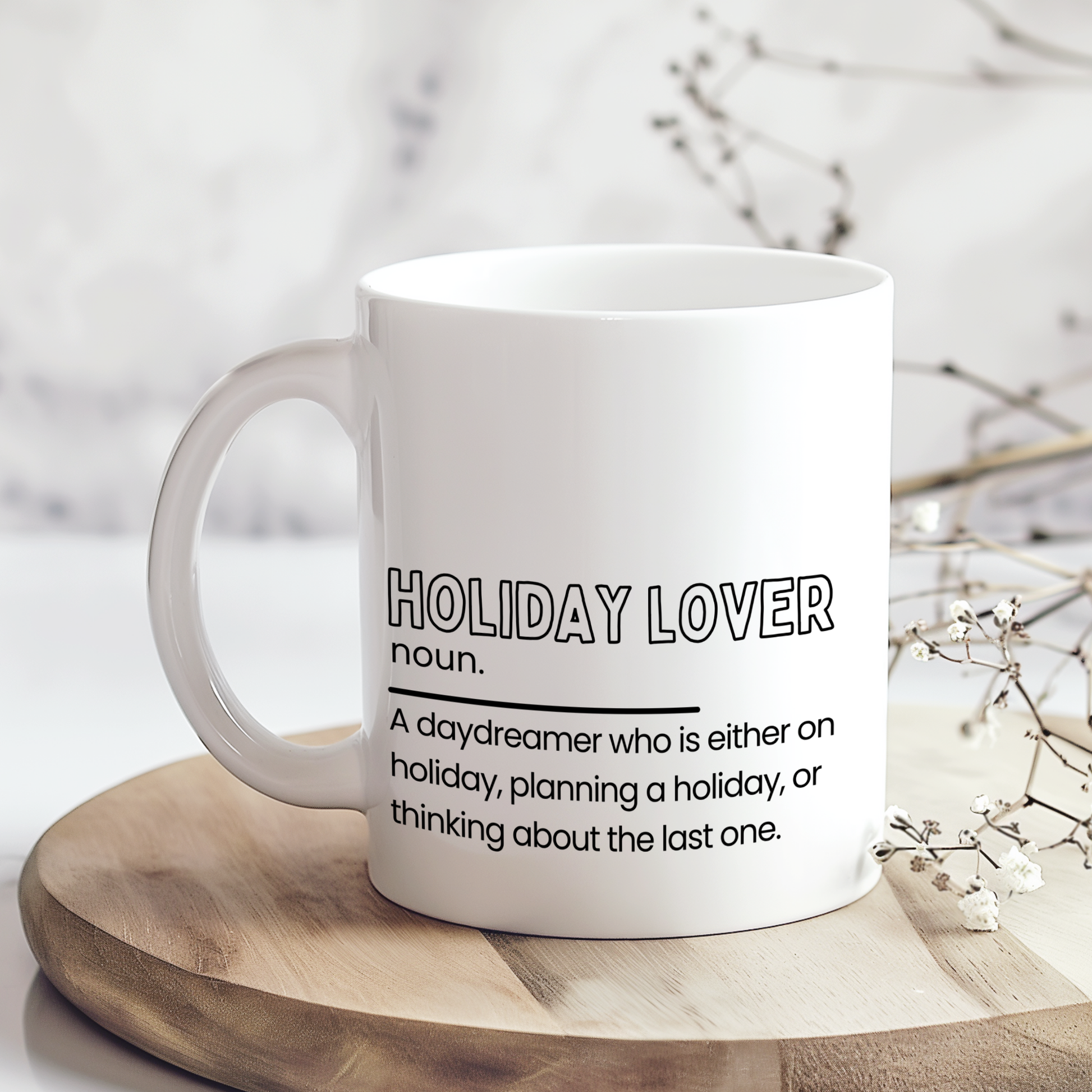 White ceramic mug with “Holiday Lover noun.” definition style quote in black typography, featuring a funny description about always being on holiday, planning one or thinking about the last trip, displayed on a neutral wooden background.