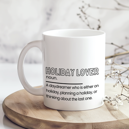 White ceramic mug with “Holiday Lover noun.” definition style quote in black typography, featuring a funny description about always being on holiday, planning one or thinking about the last trip, displayed on a neutral wooden background.