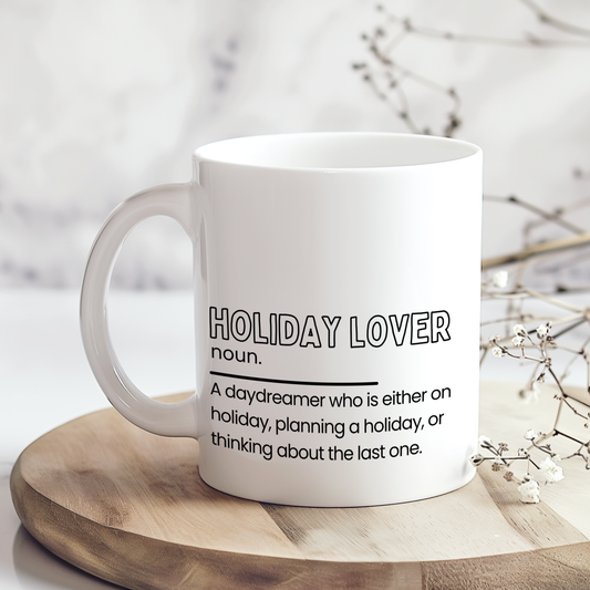 White ceramic mug with “Holiday Lover noun.” definition style quote in black typography, featuring a funny description about always being on holiday, planning one or thinking about the last trip, displayed on a neutral wooden background.