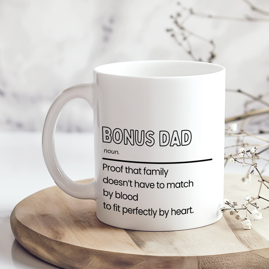 White ceramic mug with “Bonus Dad noun.” definition style quote in black typography, featuring a heartfelt message about family not needing to match by blood, shown in a lifestyle hand-held mug mockup.