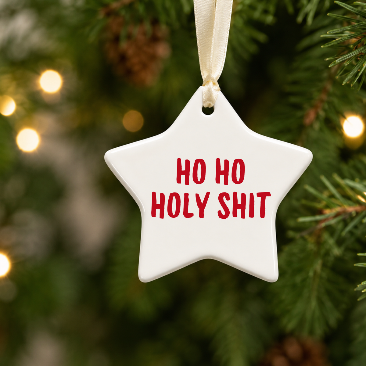 White star Christmas decoration with red “Ho Ho Holy Shit” lettering and cream ribbon, hanging on a Christmas tree with warm fairy lights.