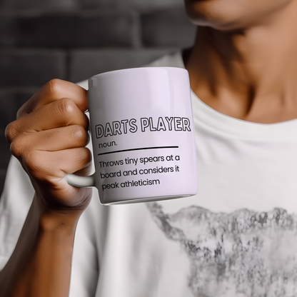 White ceramic mug featuring a humorous "Darts Player" definition with the slogan "Throws tiny spears at a board and considers it peak athleticism", making a funny gift for darts lovers.
