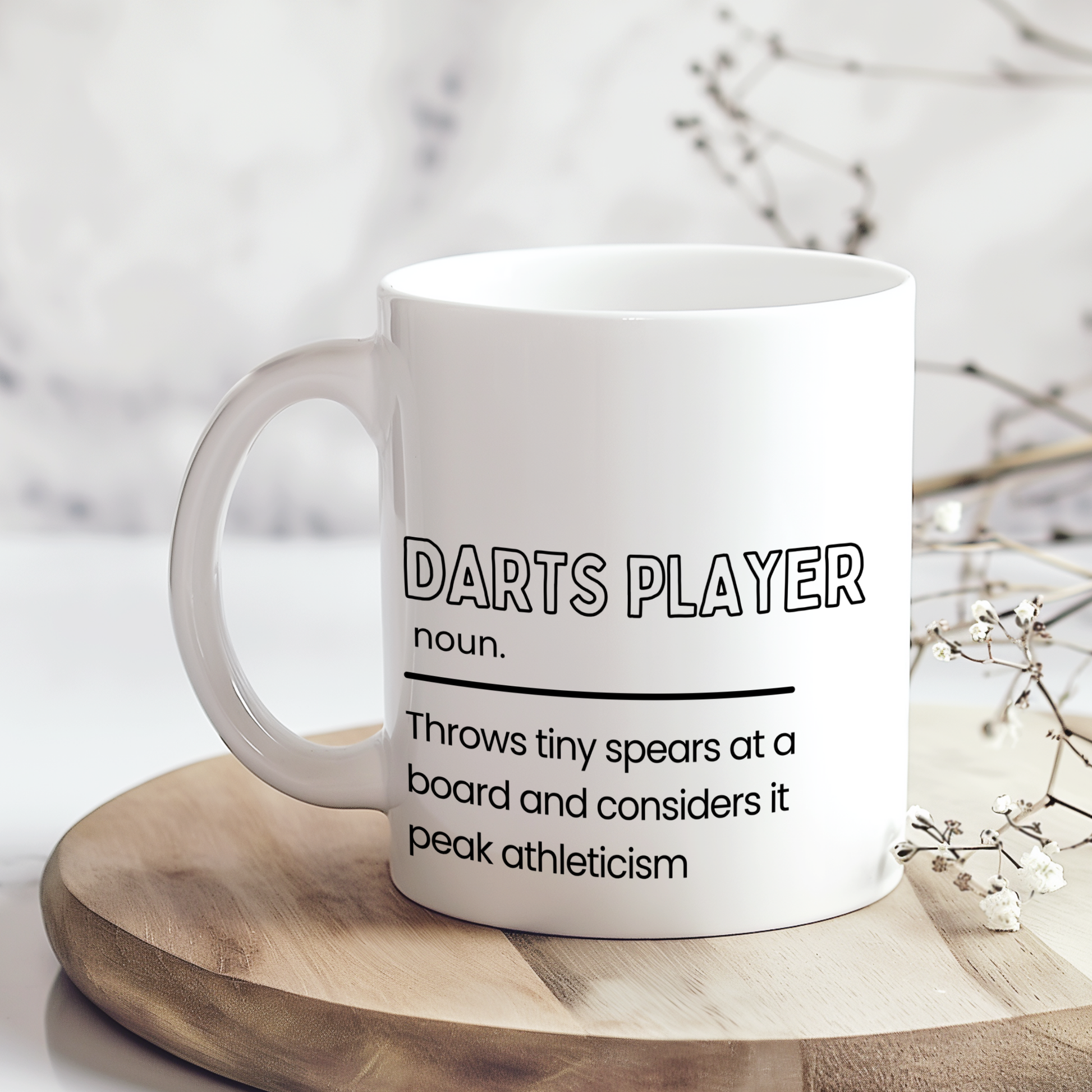 White ceramic mug featuring a humorous "Darts Player" definition with the slogan "Throws tiny spears at a board and considers it peak athleticism", making a funny gift for darts lovers.