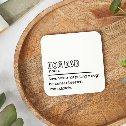 White square coaster with funny dog dad quote in black text placed on a wooden tray, humorous coaster gift for dog lovers and pet owners with a minimalist black and white design.