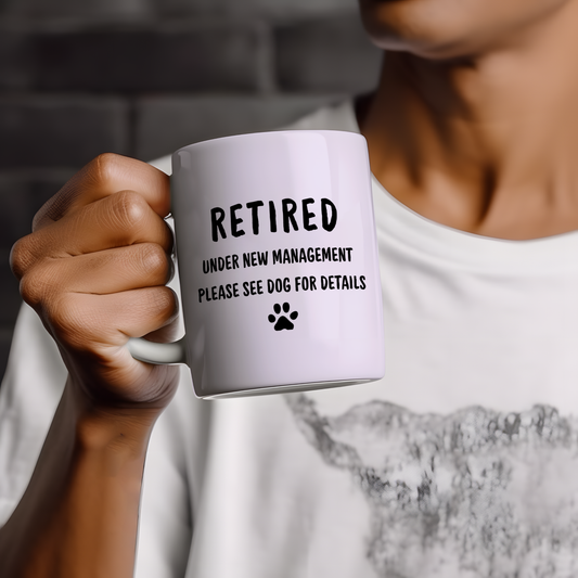 White ceramic mug with funny retirement quote reading “Retired under new management please see dog for details” in bold black typography with paw print icon, displayed on a neutral wooden background.