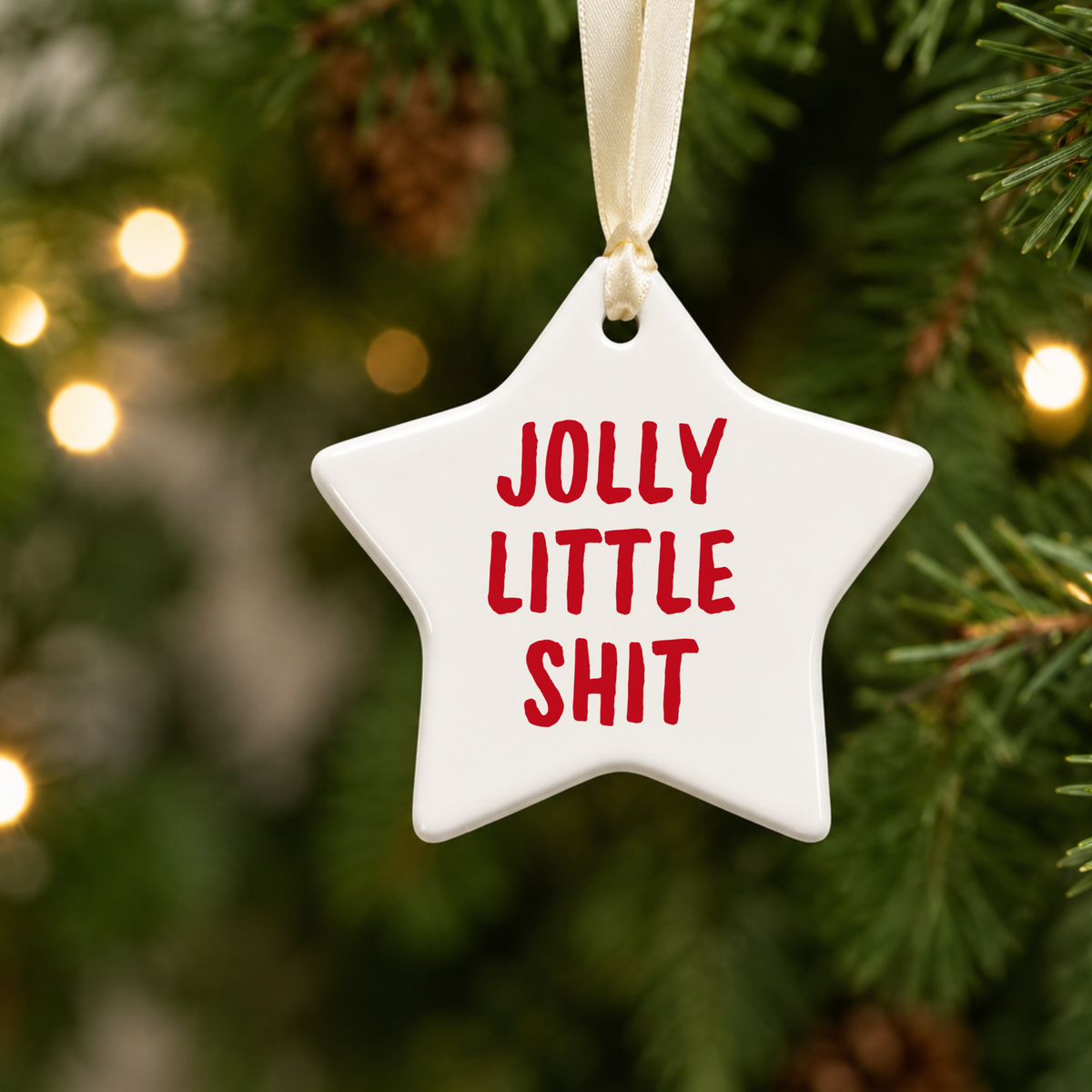 	White star Christmas decoration with red “Jolly Little Shit” lettering and cream ribbon, hanging among green Christmas tree branches and fairy lights.