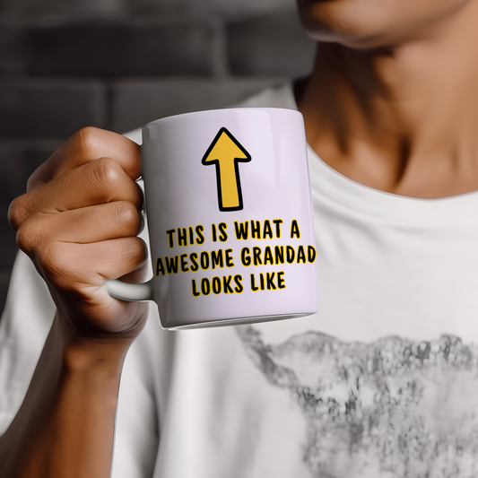 White ceramic mug with humorous “This is what an awesome grandad looks like” quote and yellow arrow graphic, shown in a lifestyle hand-held mug mockup.