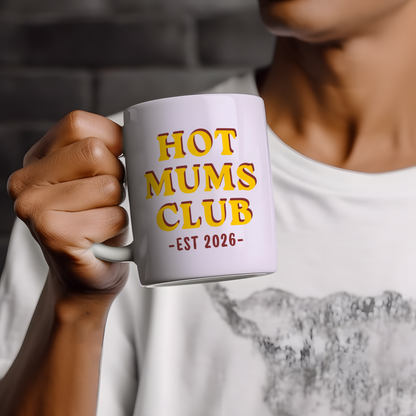 White ceramic mug featuring “Hot Mums Club EST 2026” retro style typography in yellow and red colours, displayed on a neutral wooden background with minimalist styling.