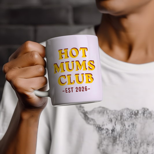 White ceramic mug featuring “Hot Mums Club EST 2026” retro style typography in yellow and red colours, displayed on a neutral wooden background with minimalist styling.