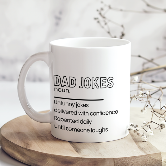 White ceramic mug with “Dad Jokes noun.” definition style design in black typography, featuring a funny description about unfunny jokes repeated daily until someone laughs, styled on a neutral wooden surface background.