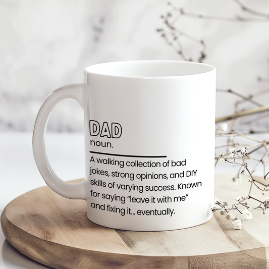 White ceramic mug featuring a funny “Dad noun.” definition quote in black typography about bad jokes, DIY skills and fixing things eventually, displayed on a neutral styled wooden surface background.