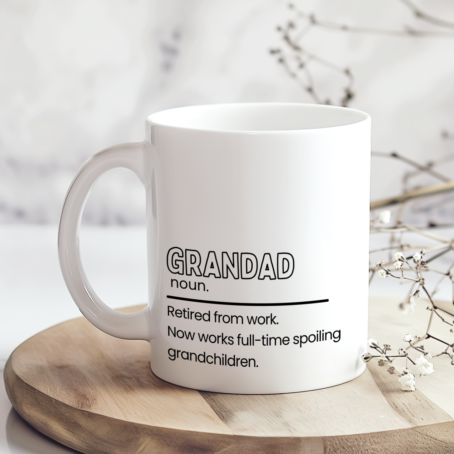 White ceramic mug with funny “Grandad” definition quote reading “Retired from work. Now works full-time spoiling grandchildren,” displayed on wooden board in neutral styled mockup, thoughtful gift mug for grandad or grandpa.
