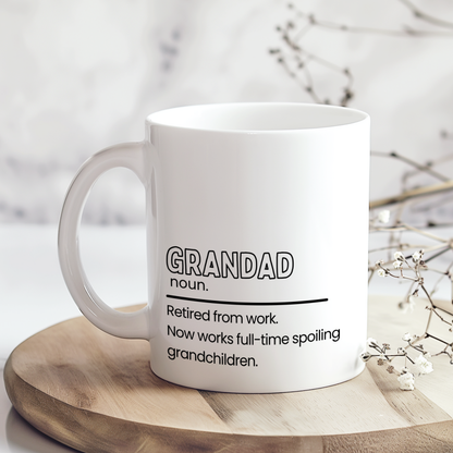White ceramic mug with funny “Grandad” definition quote reading “Retired from work. Now works full-time spoiling grandchildren,” displayed on wooden board in neutral styled mockup, thoughtful gift mug for grandad or grandpa.