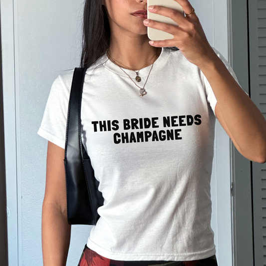 White fitted baby tee featuring the slogan “This Bride Needs Champagne” in bridal-style typography, perfect for hen party or bachelorette outfit.