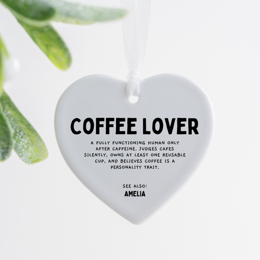 white ceramic heart ornament with coffee lover definition text hanging with white ribbon on wooden tray with pink rose