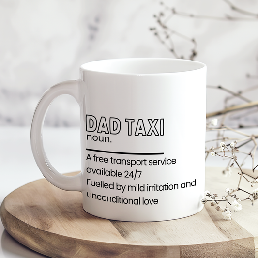 White ceramic mug with “Dad Taxi noun.” definition style quote in black typography, featuring a funny description about dads being a free transport service available 24/7, displayed on a neutral wooden backdrop.