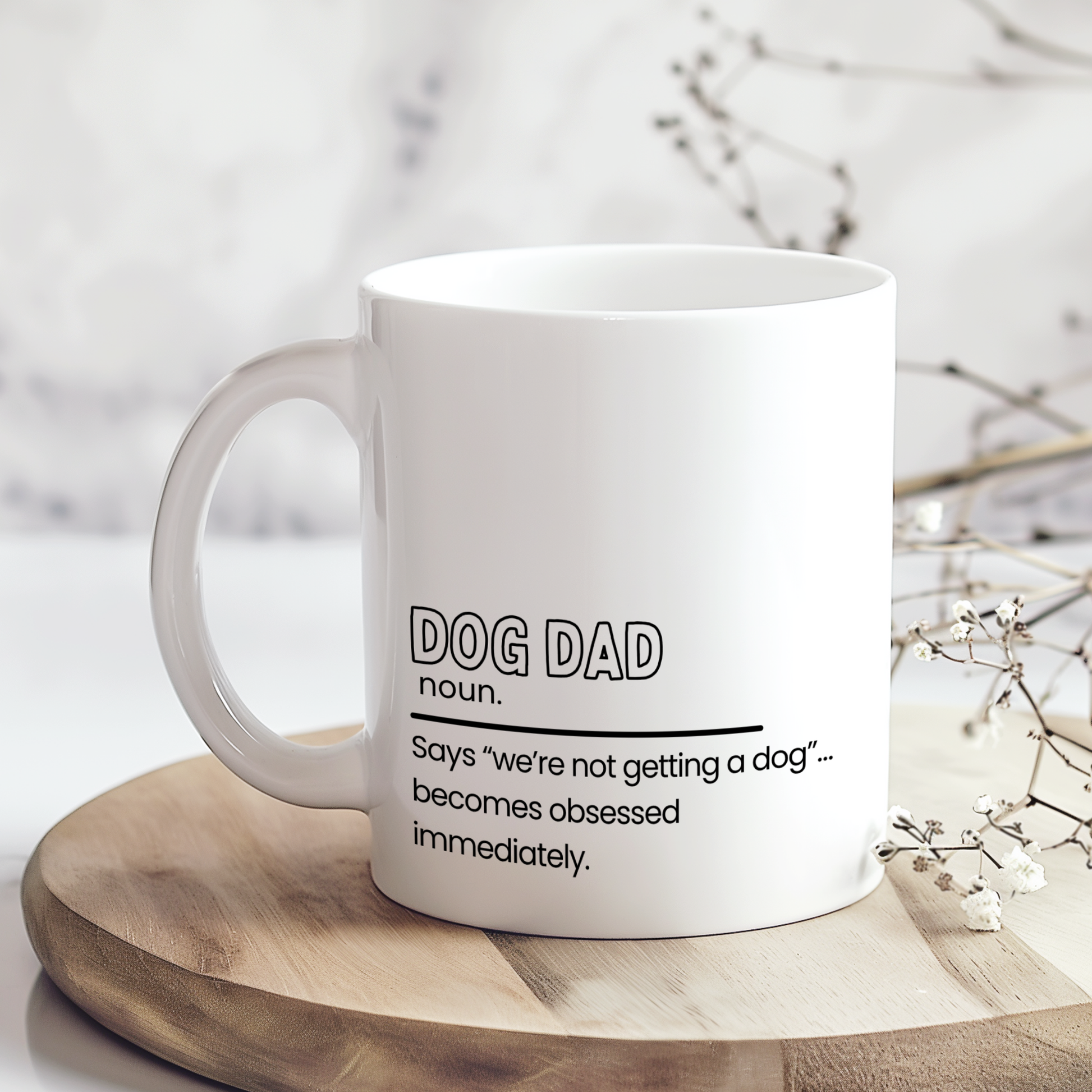 White ceramic mug with funny “Dog Dad” definition quote reading “Says ‘we’re not getting a dog’… becomes obsessed immediately,” displayed on wooden board in neutral styled mockup, humorous gift mug for dog dads and pet lovers.