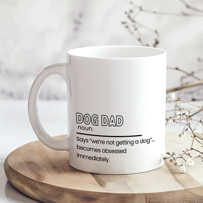 White ceramic mug with funny “Dog Dad” definition quote reading “Says ‘we’re not getting a dog’… becomes obsessed immediately,” displayed on wooden board in neutral styled mockup, humorous gift mug for dog dads and pet lovers.