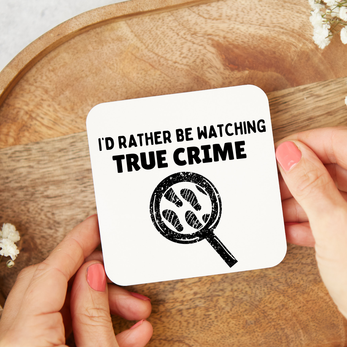 White square coaster with black “I’d Rather Be Watching True Crime” lettering and a magnifying glass containing footprints, displayed on a wooden tray.