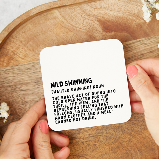 Coaster with definition of 'wild swimming' held by hands on a wooden surface