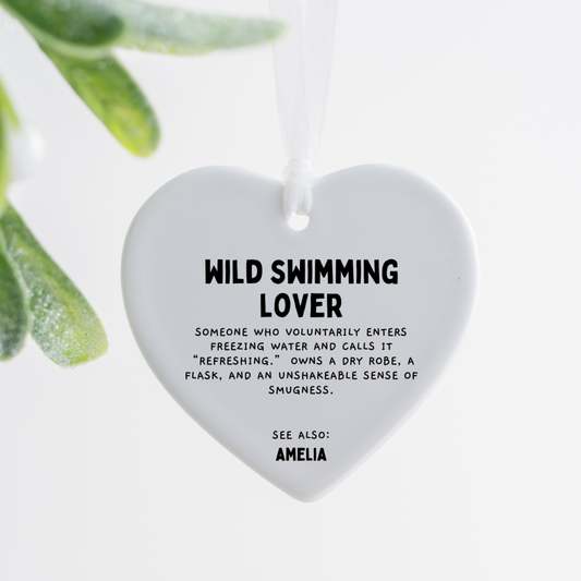 white ceramic heart ornament with wild swimming lover definition text hanging with white ribbon on wooden tray beside pink rose