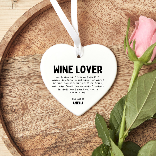 white ceramic heart ornament with wine lover definition text hanging with white ribbon on wooden tray beside pink rose